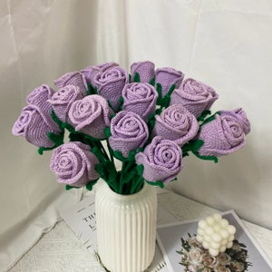 May include: A bouquet of lavender crocheted roses in a white vase. The roses are arranged in a circular pattern and have a soft, fuzzy texture.