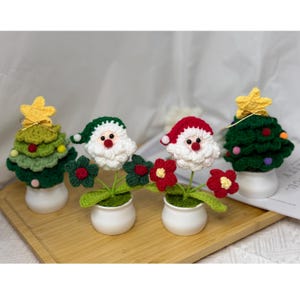 May include: A collection of handmade crochet Christmas decorations. The set includes two Christmas trees with yellow star toppers, and two Santa Claus figures with red hats. Each figure is in a small white pot, with green leaves and red flowers.