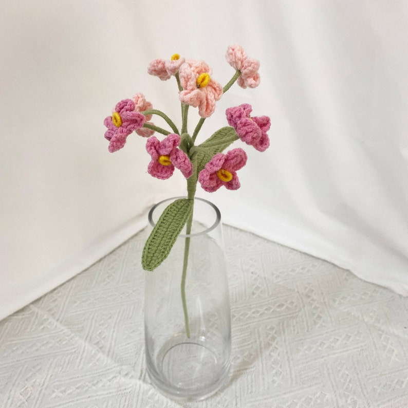 May include: A crocheted flower arrangement in a clear glass vase. The flowers are a mix of pink and light pink, with yellow centers. The stems and leaves are green. The vase sits on a white surface.