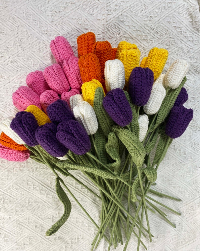 May include: A bouquet of crocheted tulips in various colors, including purple, pink, white, orange, and yellow. The tulips have green stems and leaves.