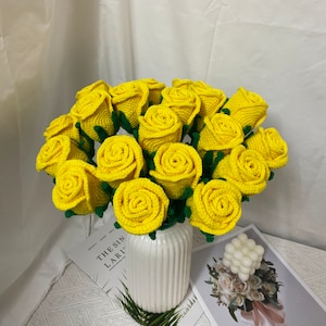 May include: A bouquet of crocheted yellow roses with green stems. The roses are arranged in a white vase with a ribbed texture. The vase is sitting on a table with a book and a small white decorative object.