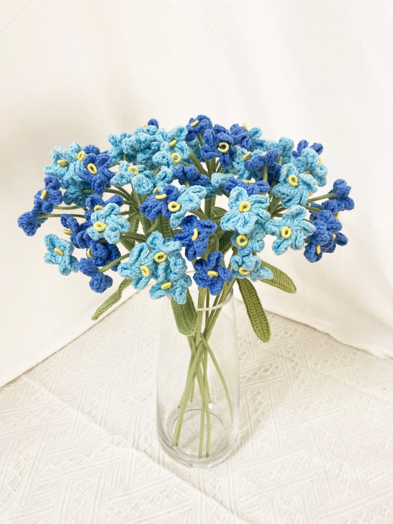 May include: A bouquet of crocheted flowers in a clear glass vase. The flowers are in shades of blue with yellow centers and green stems and leaves. The flowers are arranged in a dense cluster.