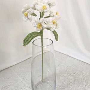 May include: A handmade crochet flower stem with white blossoms and yellow centers, green leaves, and a green stem, displayed in a clear glass vase. The vase is empty and sits on a white patterned surface.