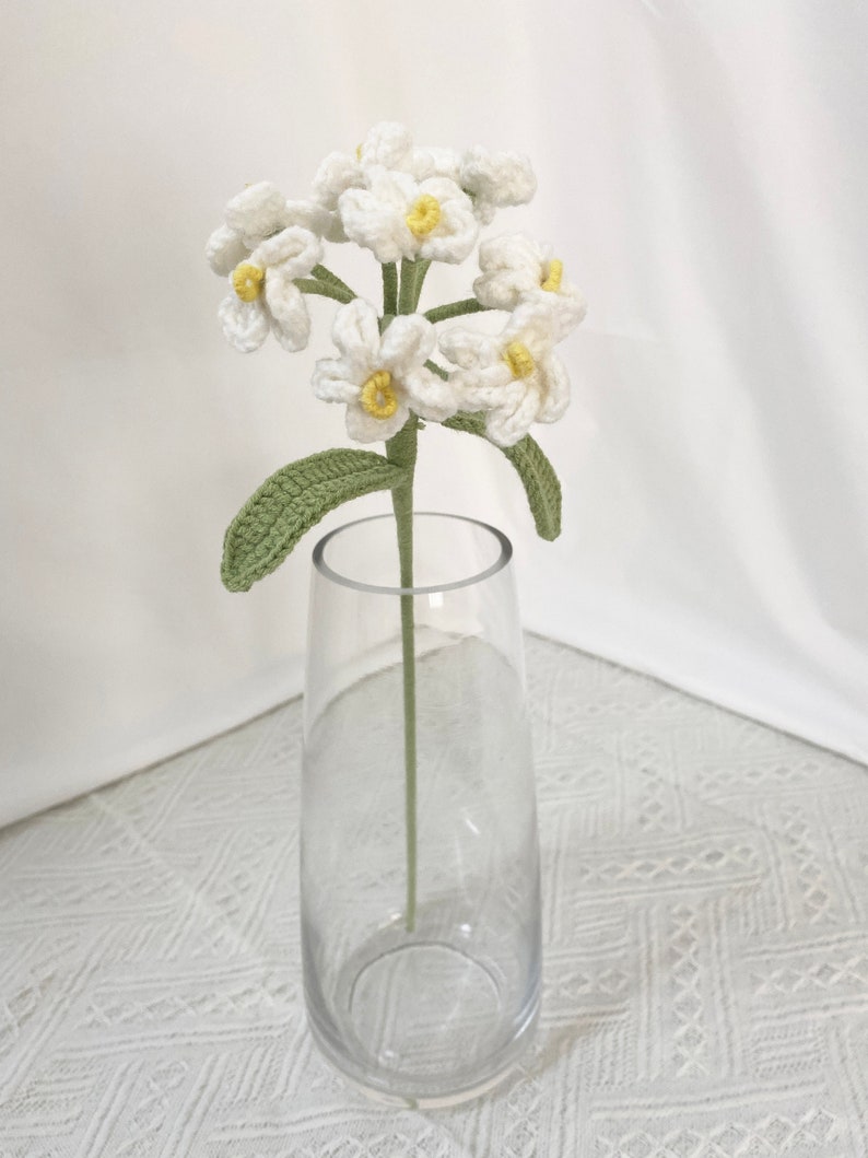 May include: A handmade crochet flower stem with white blossoms and yellow centers, green leaves, and a green stem, displayed in a clear glass vase. The vase is empty and sits on a white patterned surface.