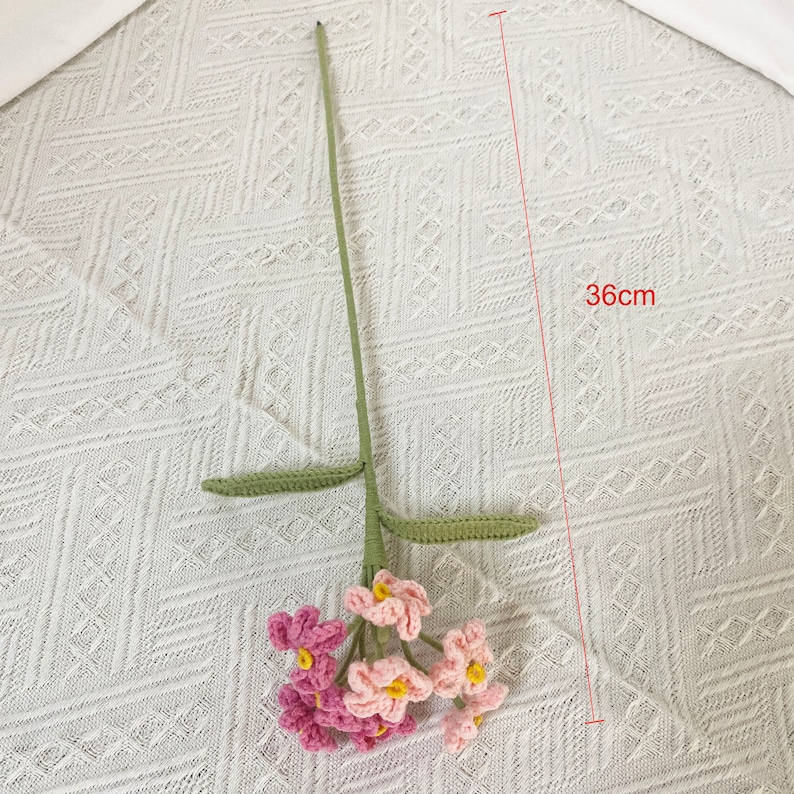 May include: A handmade crochet flower stem, approximately 14 inches long, featuring pink and light pink flowers with yellow centers. The stem and leaves are green. The flower is a decorative item.