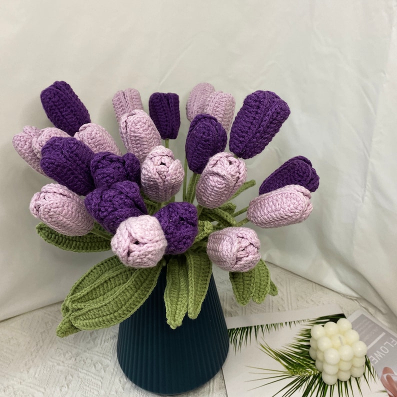 May include: A bouquet of crocheted purple and pink tulips in a dark blue vase. The tulips are arranged in a vase with green crocheted leaves.