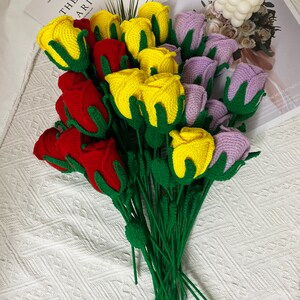 May include: A bouquet of crocheted roses in red, yellow, and purple. The roses are arranged in a fan shape and have green stems.