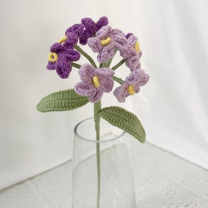 May include: A handmade crochet flower with purple and lavender petals and yellow centers. The flower has a green stem and leaves. The flower is displayed in a clear glass vase.