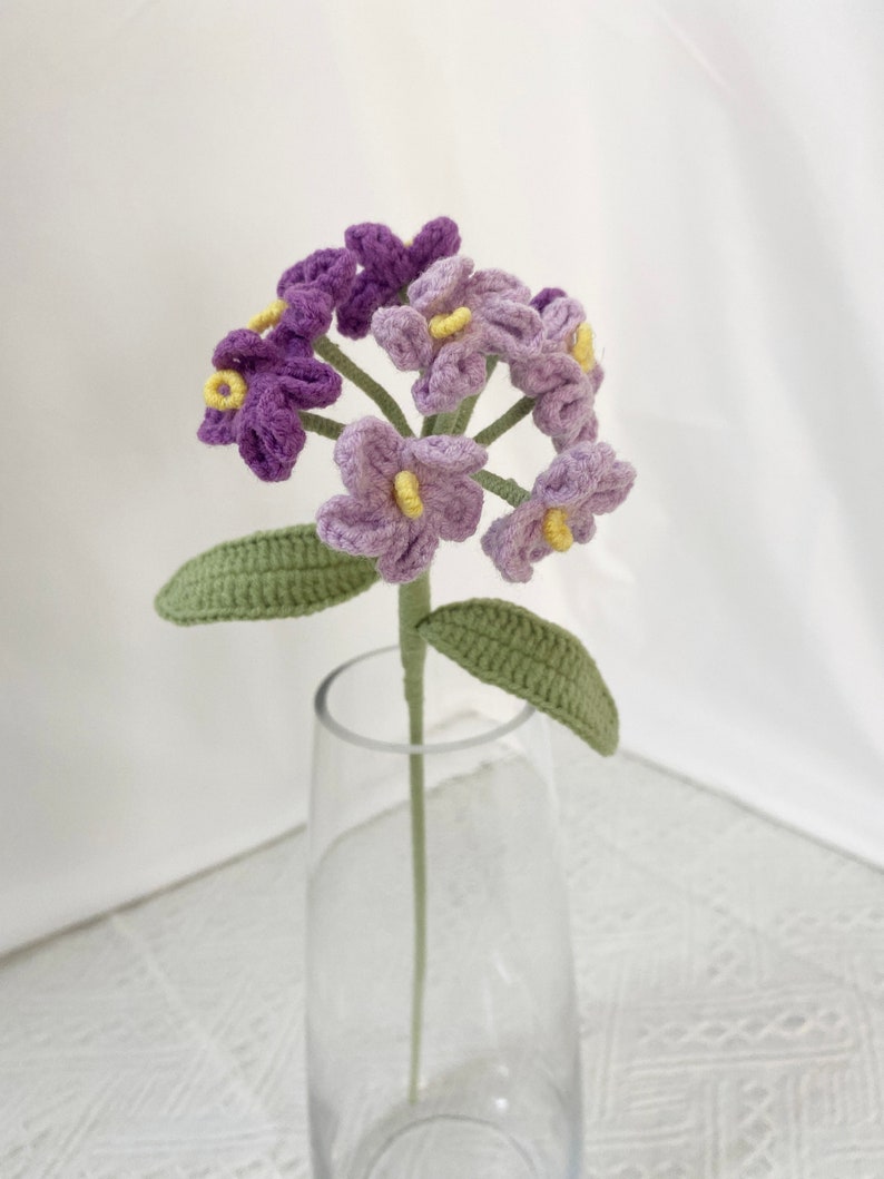 May include: A handmade crochet flower with purple and lavender petals and yellow centers. The flower has a green stem and leaves. The flower is displayed in a clear glass vase.