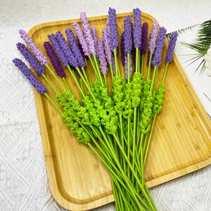 May include: A bouquet of crocheted lavender flowers with green stems. The flowers are arranged on a wooden tray.