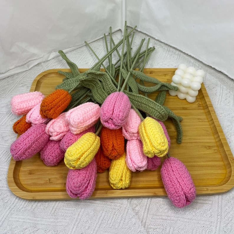 May include: A bouquet of crocheted tulips in pink, yellow, and orange. The tulips are arranged on a wooden tray.