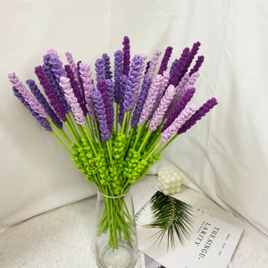 May include: A bouquet of purple and green crocheted lavender flowers in a clear glass vase. The vase is sitting on a table with a book and a white decorative object.