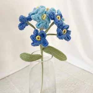 May include: A handmade crochet flower arrangement featuring blue and light blue forget-me-nots with yellow centers. The flowers are displayed in a clear glass vase, with green leaves and stems. A unique, handcrafted floral home decor item.