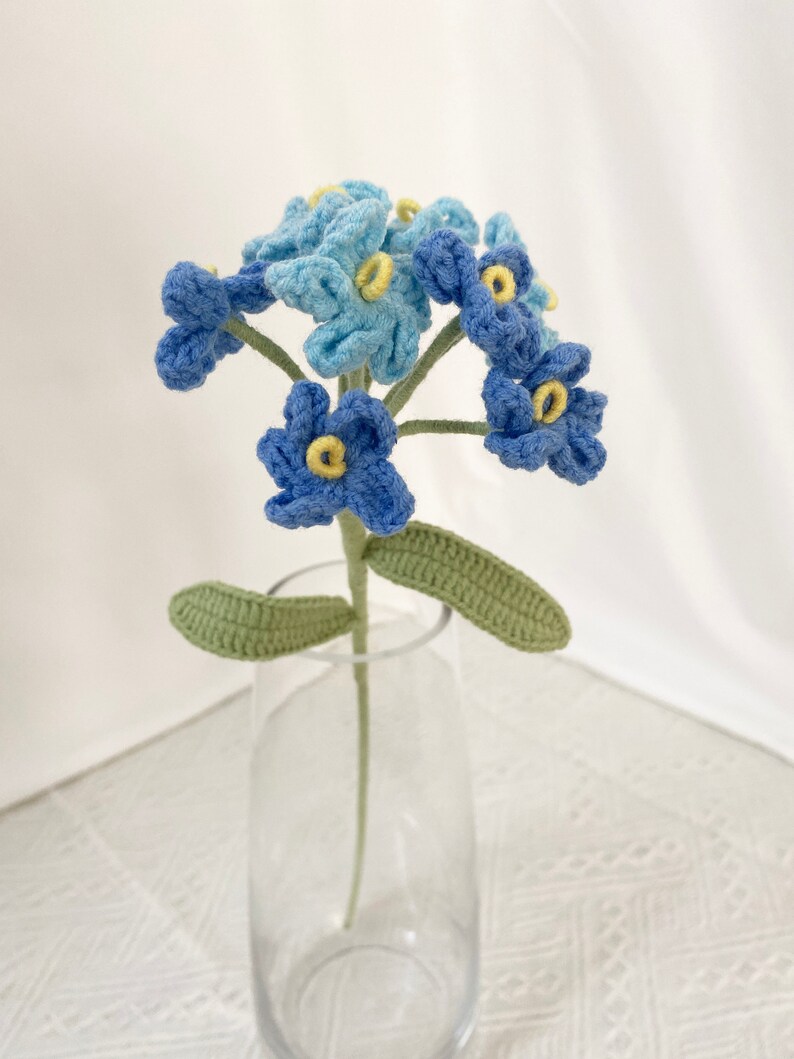 May include: A handmade crochet flower arrangement featuring blue and light blue forget-me-nots with yellow centers. The flowers are displayed in a clear glass vase, with green leaves and stems. A unique, handcrafted floral home decor item.