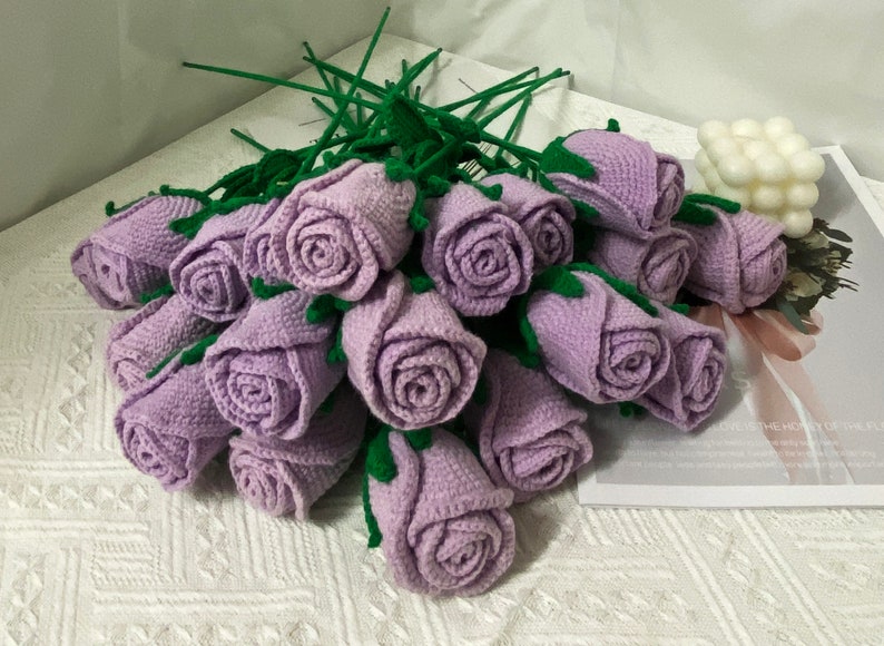 May include: A bouquet of crocheted lavender roses with green stems. The roses are arranged in a circular pattern and are tied together with a green ribbon.