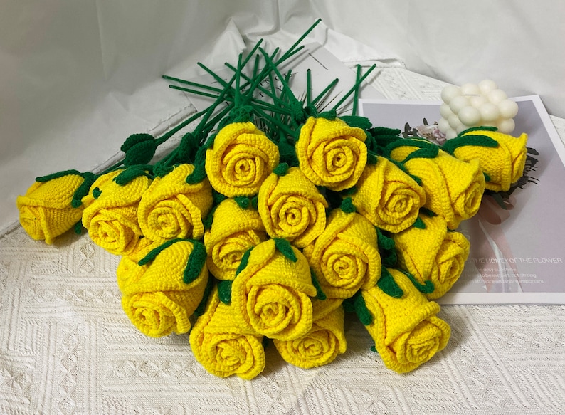 May include: A bouquet of crocheted yellow roses with green leaves and stems. The roses are arranged in a circular shape, with the stems extending outwards.