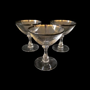 May include: Three clear glass champagne coupes with a silver rim.