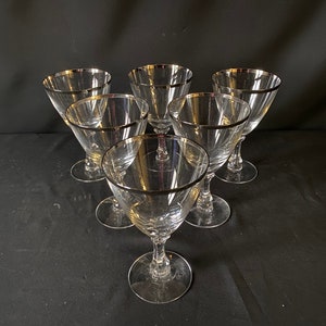 May include: Set of six clear glass wine glasses with a silver rim. The glasses are arranged in a circle on a black surface.