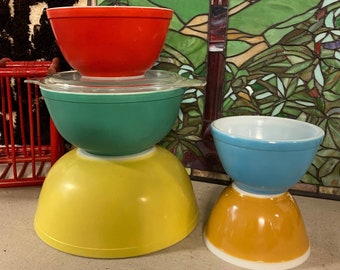 May include: Four vintage mixing bowls stacked in a pyramid shape. The top bowl is red, the second bowl is green, the third bowl is yellow, and the bottom bowl is blue with an orange base.