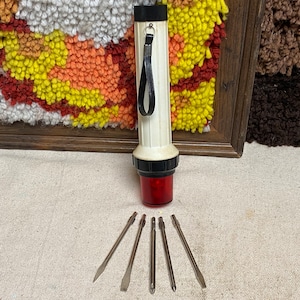 May include: A vintage flashlight with a cream-colored body, black accents, and a red lens. Five screwdrivers are arranged below the flashlight. The background includes a framed, colorful textile art piece.