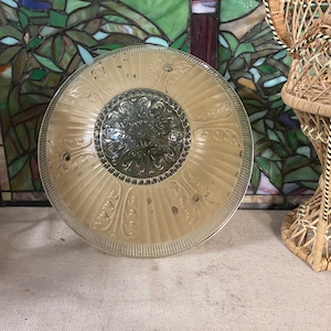 May include: A vintage, round, glass light fixture with a cream-colored, ribbed design and a decorative center. The fixture has a clear, etched edge and several small holes. The background includes a stained glass window and a wicker chair.