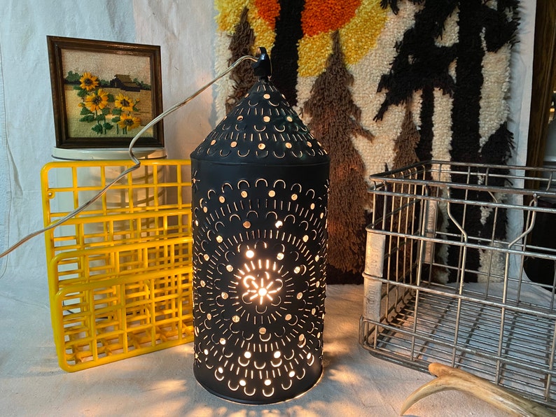 Puede incluir: Una linterna de metal negro con un dise&ntilde;o recortado y una luz en su interior. La linterna est&aacute; sentada sobre una superficie blanca con una rejilla de alambre amarilla y una alfombra marr&oacute;n y blanca en el fondo.