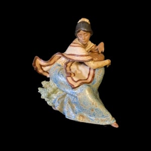 May include: A porcelain figurine of a woman in a blue and brown dress, holding a pitcher. She is wearing a white shawl and has brown hair.