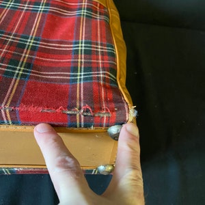 Op de afbeelding: Close-up van de onderkant van een vintage koffer met een bruine leren buitenkant en een rode en zwarte geruite voering. De koffer heeft metalen studs langs de rand.