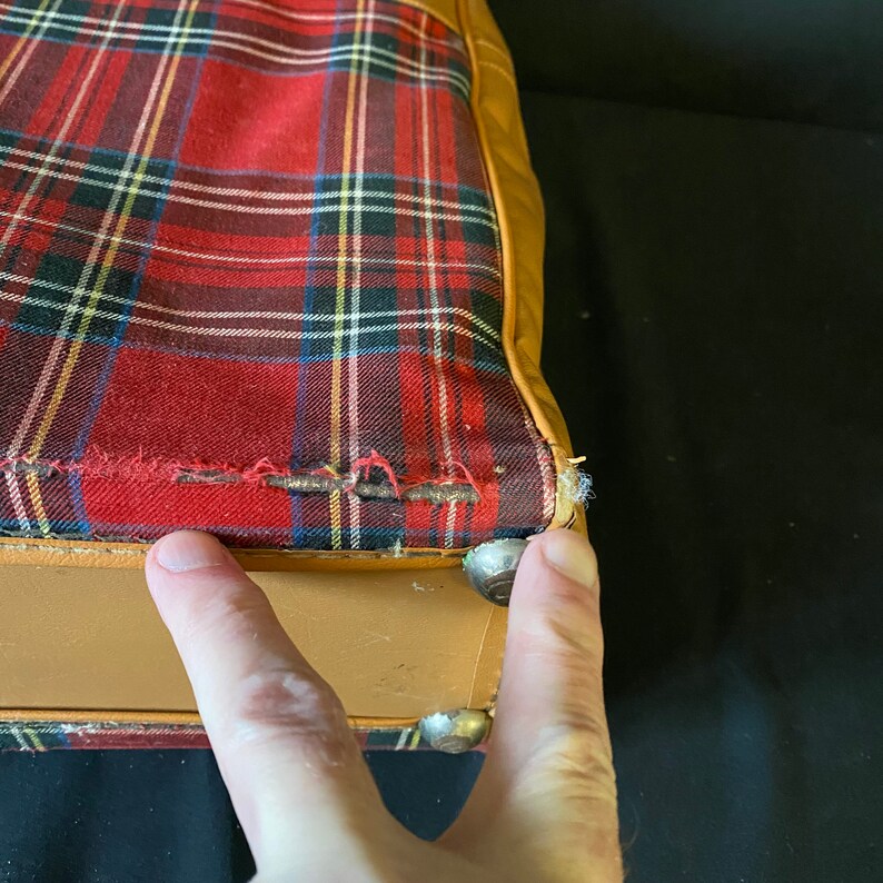 Op de afbeelding: Close-up van de onderkant van een vintage koffer met een bruine leren buitenkant en een rode en zwarte geruite voering. De koffer heeft metalen studs langs de rand.