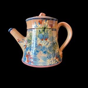 May include: A hand-painted ceramic teapot with a floral design in shades of blue, red, and green. The teapot has a rounded body and a spout on the side.