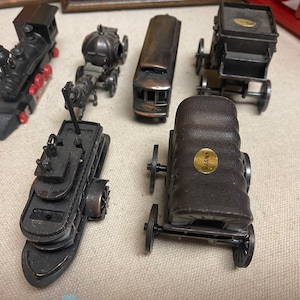 May include: A collection of six vintage metal pencil sharpeners shaped like a train, a carriage, a boat, a bus, and two covered wagons. The sharpeners are all made of dark metal and have a worn, antique look.