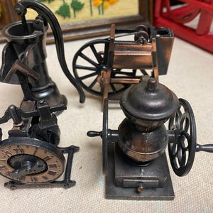 May include: Three vintage-style metal pencil sharpeners. One is a well pump, one is a coffee grinder, and one is a wagon. All are made of metal and have a bronze finish.