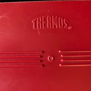 Op de afbeelding: Rode thermoskan met het woord "THERMOS" in reli&euml;f op de bovenkant. De kan heeft vijf horizontale lijnen en een kleine cirkel in het midden.