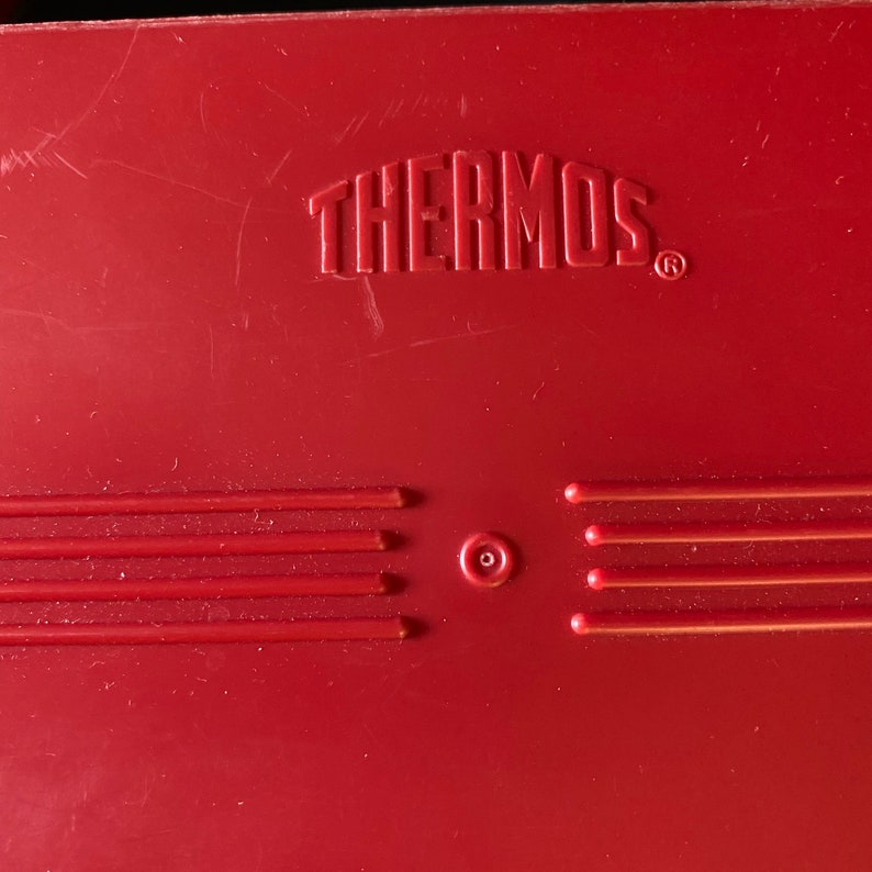 Op de afbeelding: Rode thermoskan met het woord "THERMOS" in reli&euml;f op de bovenkant. De kan heeft vijf horizontale lijnen en een kleine cirkel in het midden.