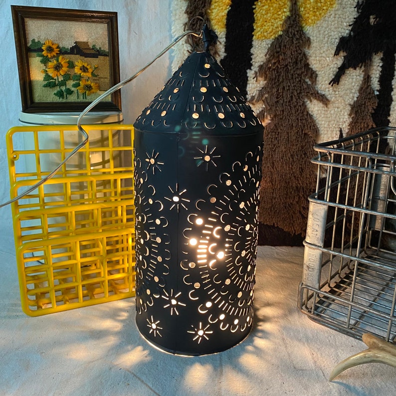 Puede incluir: Una linterna de metal negro con un dise&ntilde;o recortado de estrellas y c&iacute;rculos. La linterna est&aacute; iluminada desde adentro, proyectando un brillo c&aacute;lido.