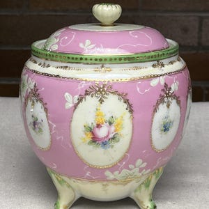 Op de afbeelding: Een roze en witte porseleinen pot met een deksel, met handgeschilderde bloemmotieven in ovale kaders. De pot heeft een groene en gouden rand, een witte knop en staat op drie kleine voetjes. Een decoratief stuk.