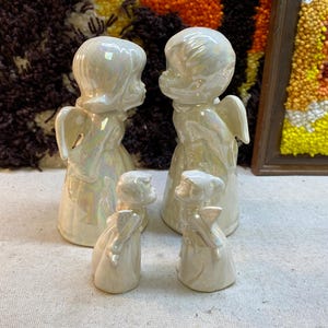 May include: Four white porcelain angel figurines. Two larger angels are standing with their wings spread out. Two smaller angels are standing with their wings folded behind them. The angels are all facing forward and have a pearlescent finish.