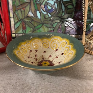 May include: A small, round, hand-painted ceramic bowl. The exterior is a muted teal, while the interior features a cream-colored base with yellow and gold decorative swirls and floral accents. The rim is trimmed with gold.