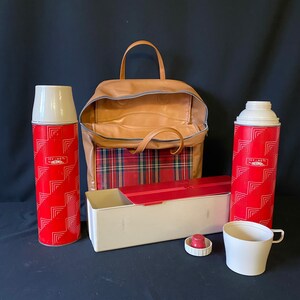 Op de afbeelding: Een vintage picknickkoeltas met een rood en zwart ruitpatroon, een bruin leren handvat en twee rode en witte thermoskannen met de tekst "Icy-Hot Thermos" erop.