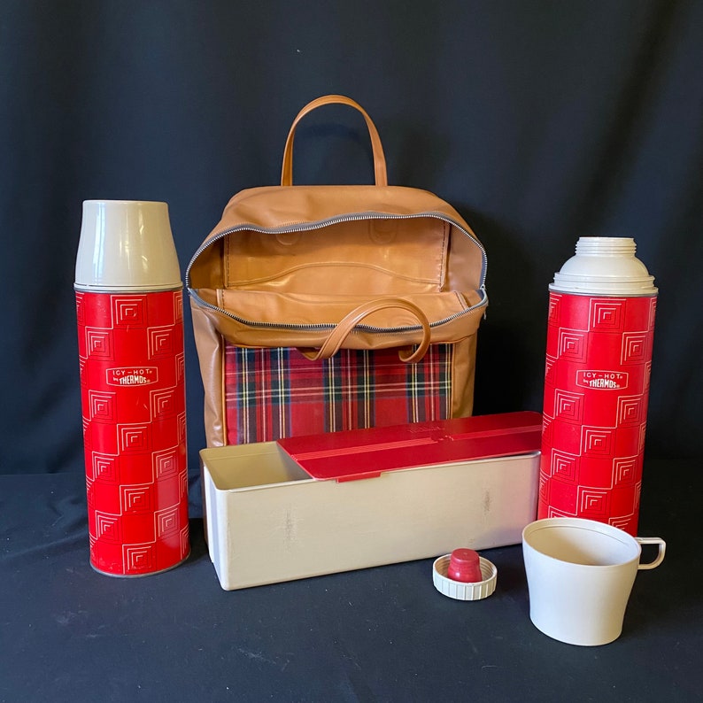 Op de afbeelding: Een vintage picknickkoeltas met een rood en zwart ruitpatroon, een bruin leren handvat en twee rode en witte thermoskannen met de tekst "Icy-Hot Thermos" erop.
