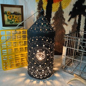 Puede incluir: Una linterna de metal negro con un dise&ntilde;o recortado y una luz en su interior. La linterna est&aacute; sentada sobre una mesa con un patr&oacute;n de cuadros amarillos y blancos.