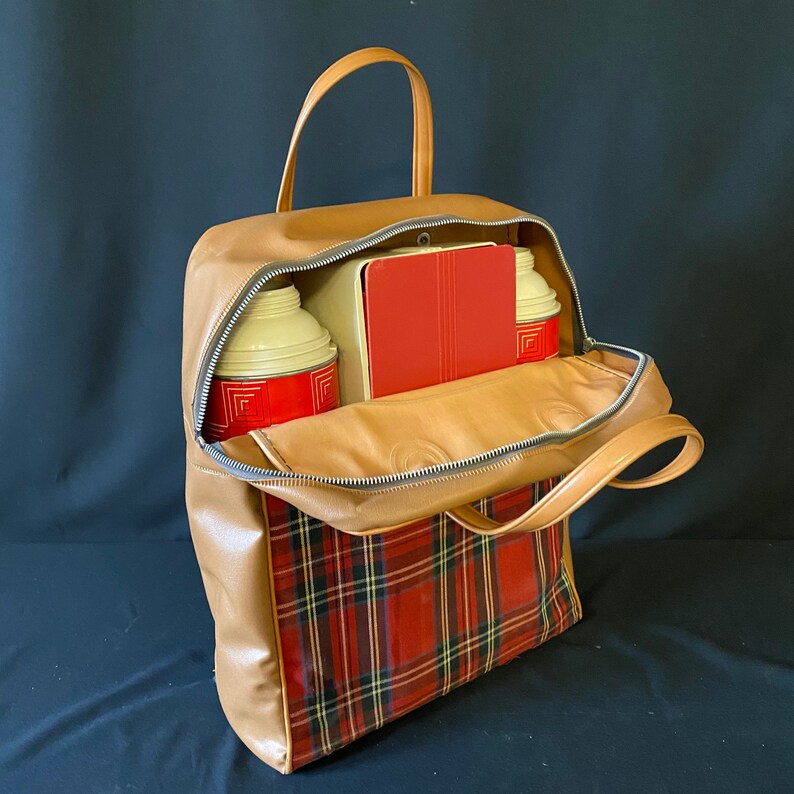 Op de afbeelding: Een bruine leren picknicktas met een rode en zwarte geruite voering. De tas is open en bevat twee metalen thermoskannen en een rode metalen lunchbox.