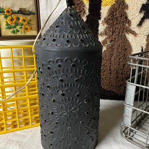 Puede incluir: Una linterna de metal negro con un dise&ntilde;o decorativo de corte. La linterna tiene una parte superior en forma de cono y un cuerpo cil&iacute;ndrico. El dise&ntilde;o de corte presenta c&iacute;rculos, flores y otros patrones.