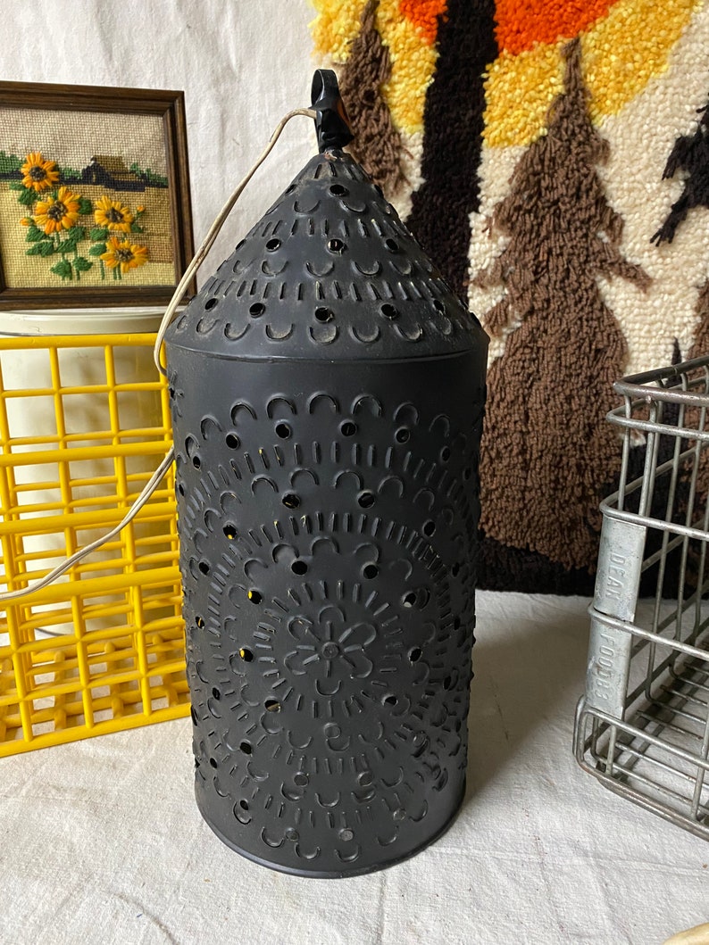 Puede incluir: Una linterna de metal negro con un dise&ntilde;o decorativo de corte. La linterna tiene una parte superior en forma de cono y un cuerpo cil&iacute;ndrico. El dise&ntilde;o de corte presenta c&iacute;rculos, flores y otros patrones.
