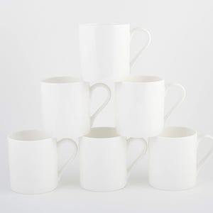 May include: Six white ceramic mugs stacked in a pyramid formation. Each mug has a curved handle. The mugs are set against a plain white backdrop. The mugs are all the same size and shape.