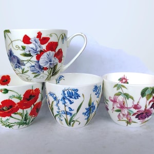 May include: A set of four white ceramic mugs featuring botanical illustrations. One mug displays red and blue sweet peas, another shows bright red poppies, the third features blue bellflowers, and the fourth depicts pink and purple fuchsia flowers.