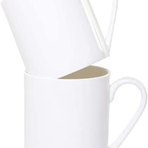 May include: Two plain white ceramic mugs stacked vertically. The top mug is tilted at an angle, while the bottom mug sits upright. Both mugs feature a smooth, glossy finish, a cylindrical body, and a curved handle attached to the side.