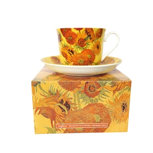 May include: A ceramic teacup and saucer set with a sunflower design, presented on a matching box. The cup and saucer have a white rim and handle. The box is yellow with a sunflower pattern. The text on the box reads "Sunflower Breakfast Cup & Saucer - For Fine Bone China."