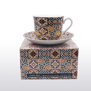 May include: A white teacup and saucer with a black and gold geometric pattern. The set is in a box with the same pattern and the text "Azulejo Black" and "Breakfast Cup & Saucer - Fine Bone China".
