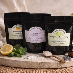 Può includere: Tre sacchetti neri di eliminatore di odori per tappeti e tappezzeria con etichette che riportano "Lemon Mint", "Lavender" e "Bergamot Rosemary". Una metà di limone, rametti di menta e una piccola bottiglia sono accanto ai sacchetti. Un cucchiaio di polvere è su una pietra.